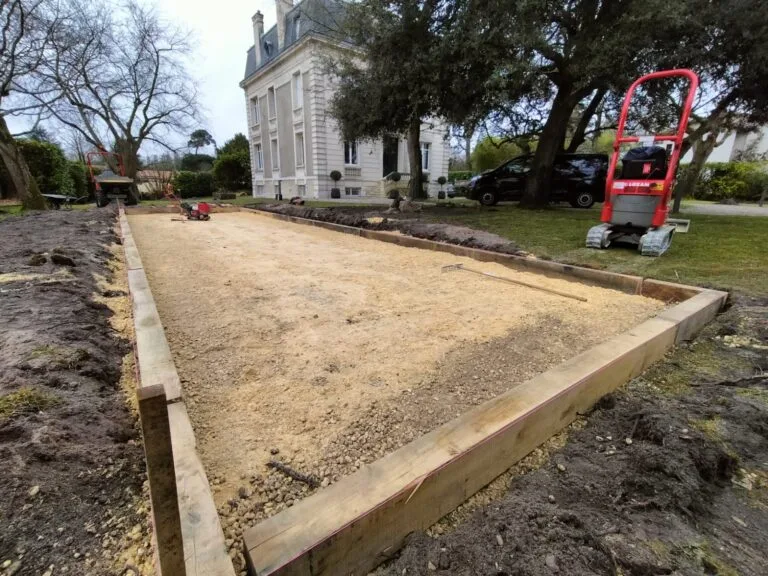 Terrain de pétanque terminé, bordé de bois, devant une maison en pierre