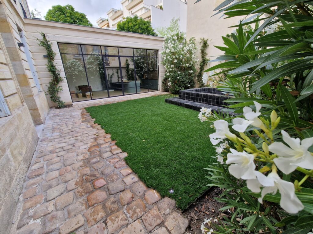 Jardin de ville terminé : gazon, allée pavée, banquette carrelée noire et laurier-rose en fleurs devant la véranda