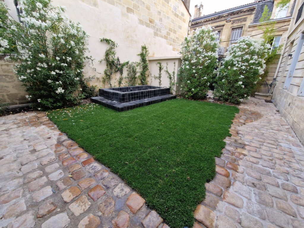 Cour aménagée : pelouse encadrée de pavés en pierre, bassin carrelé noir et rosiers grimpants
